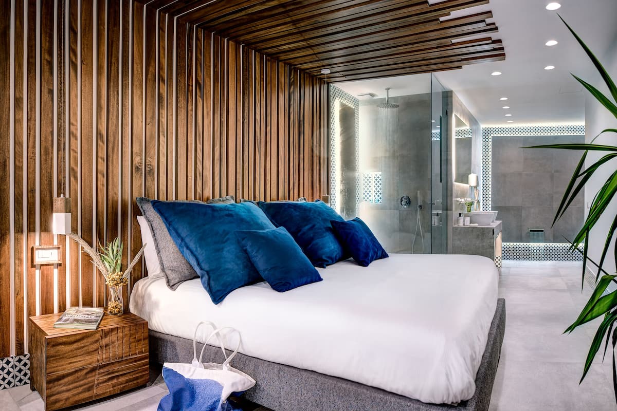 Ein modernes Schlafzimmer mit einer Wand aus Holzlamellen, einem großen Bett mit blauen Kissen, einem Nachttisch mit einer Zeitschrift und einem durch eine Glastür einsehbaren Badezimmer.