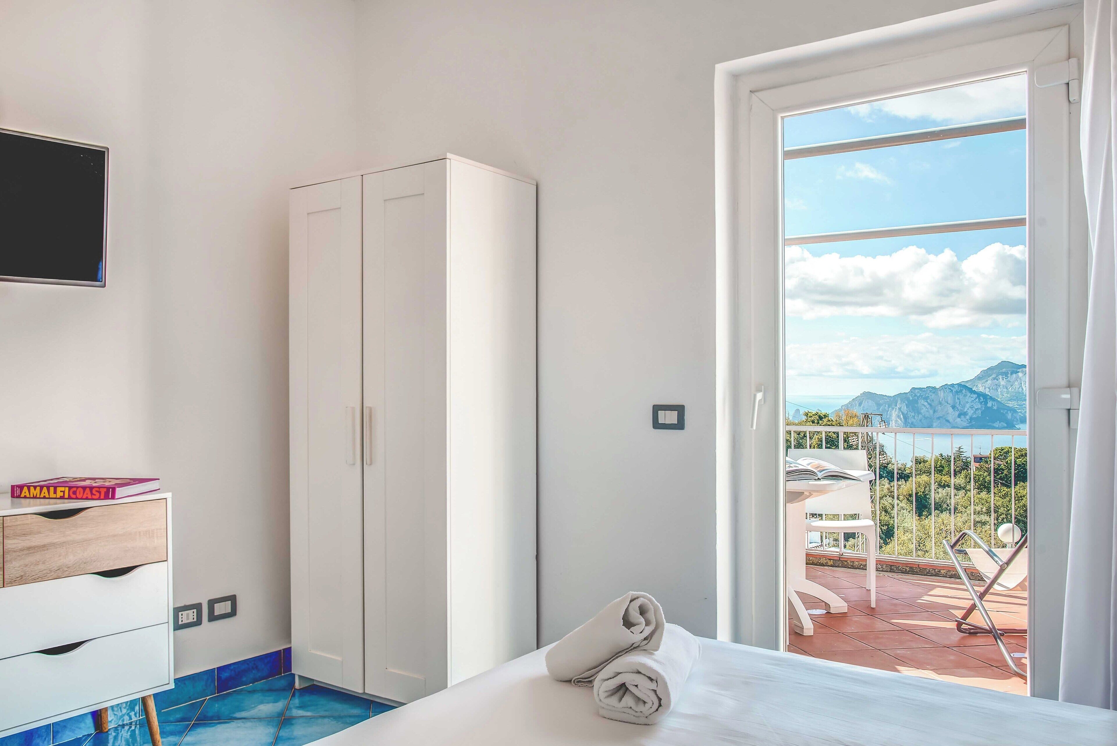 A hotel room with a bed, a wardrobe, a television, and a balcony with a view of mountains and a lake.