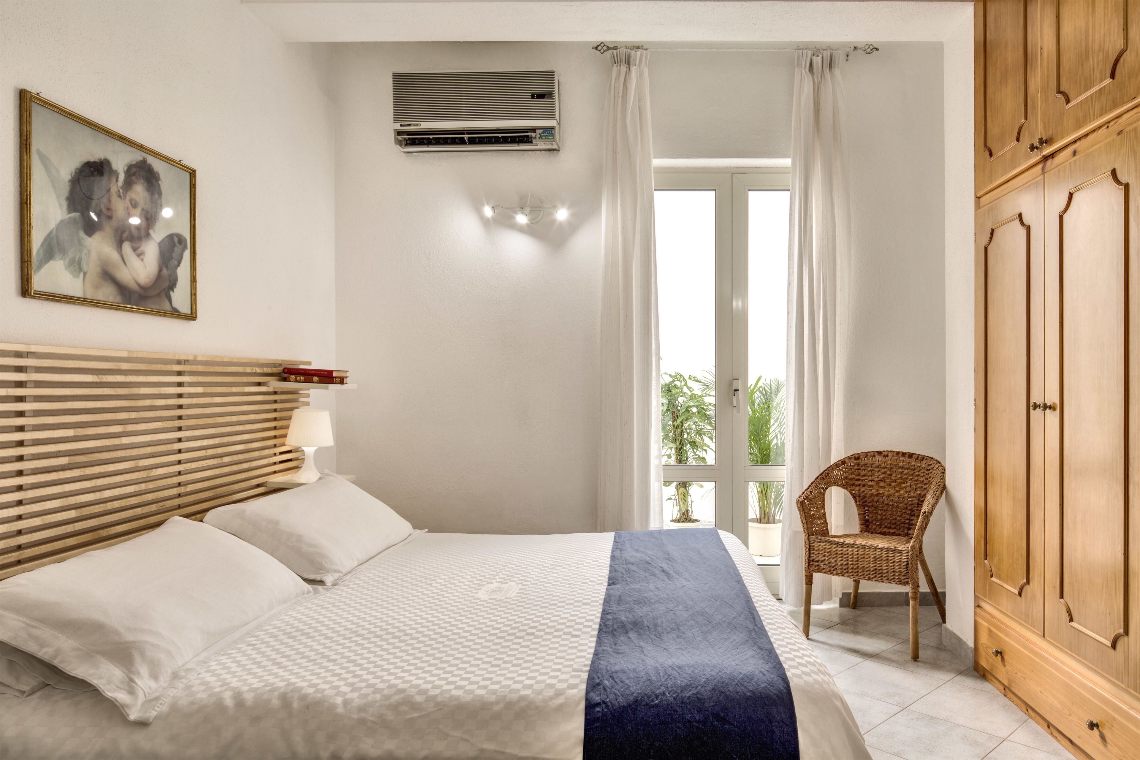 A bedroom with a bed, a wooden wardrobe, a wicker chair, and a balcony with curtains.