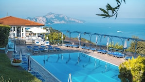A pool area with lounge chairs and umbrellas, a covered seating area, and a view of the sea and mountains.