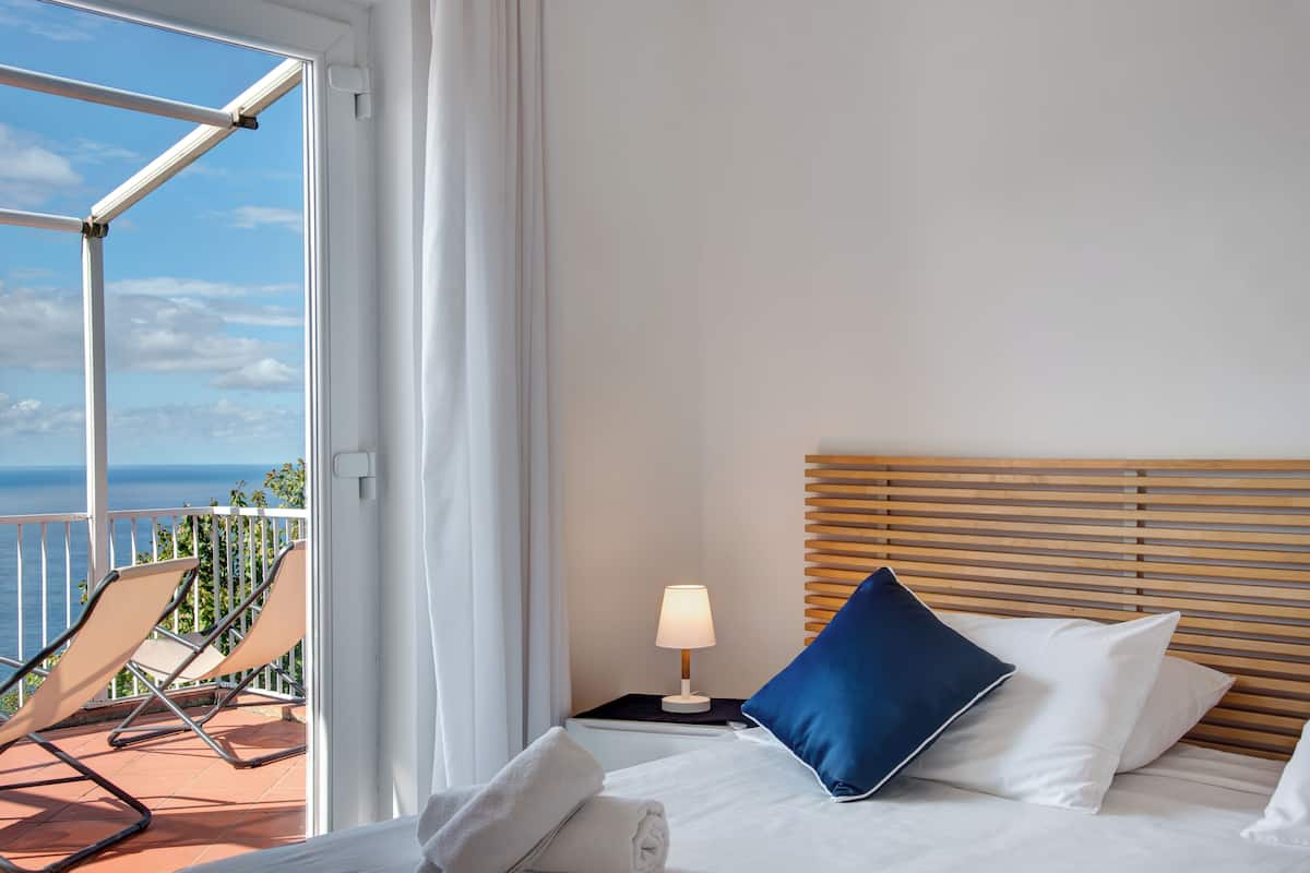 Ein Hotelzimmer mit einem Bett, einem Nachttisch mit Lampe, einem blauen Kissen und Meerblick vom Balkon.