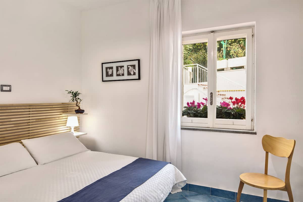 Ein Schlafzimmer mit Bett, Holz-Kopfstück, Nachttisch mit Lampe, Stuhl und Fenster mit Blick auf einen Balkon und Blumen.