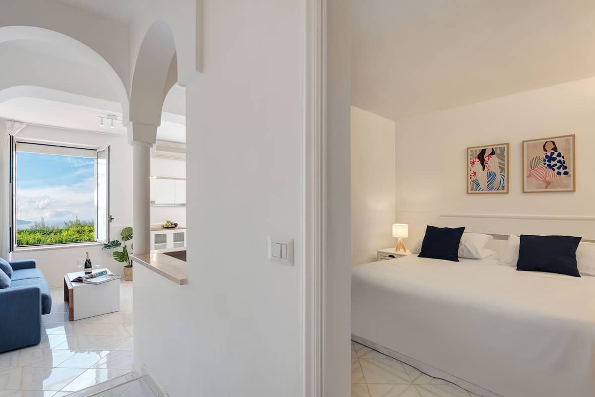 Ein modernes Hotelzimmer mit Bett, einer Sitzecke mit Sofa, einem Esstisch und Blick ins Freie.