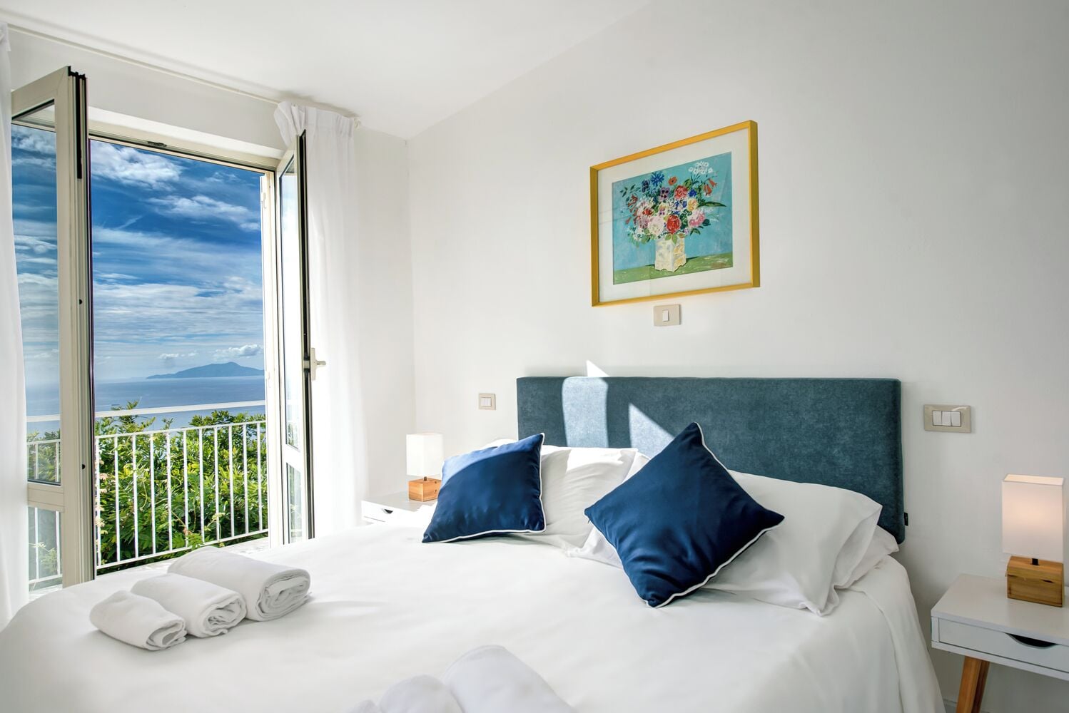 Habitación de hotel con cama, almohadas, mesita de noche, lámpara, un cuadro y un balcón con vistas.