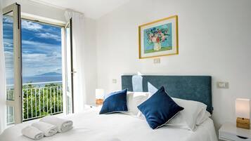 Habitación de hotel con cama, almohadas, mesita de noche, lámpara, un cuadro y un balcón con vistas.