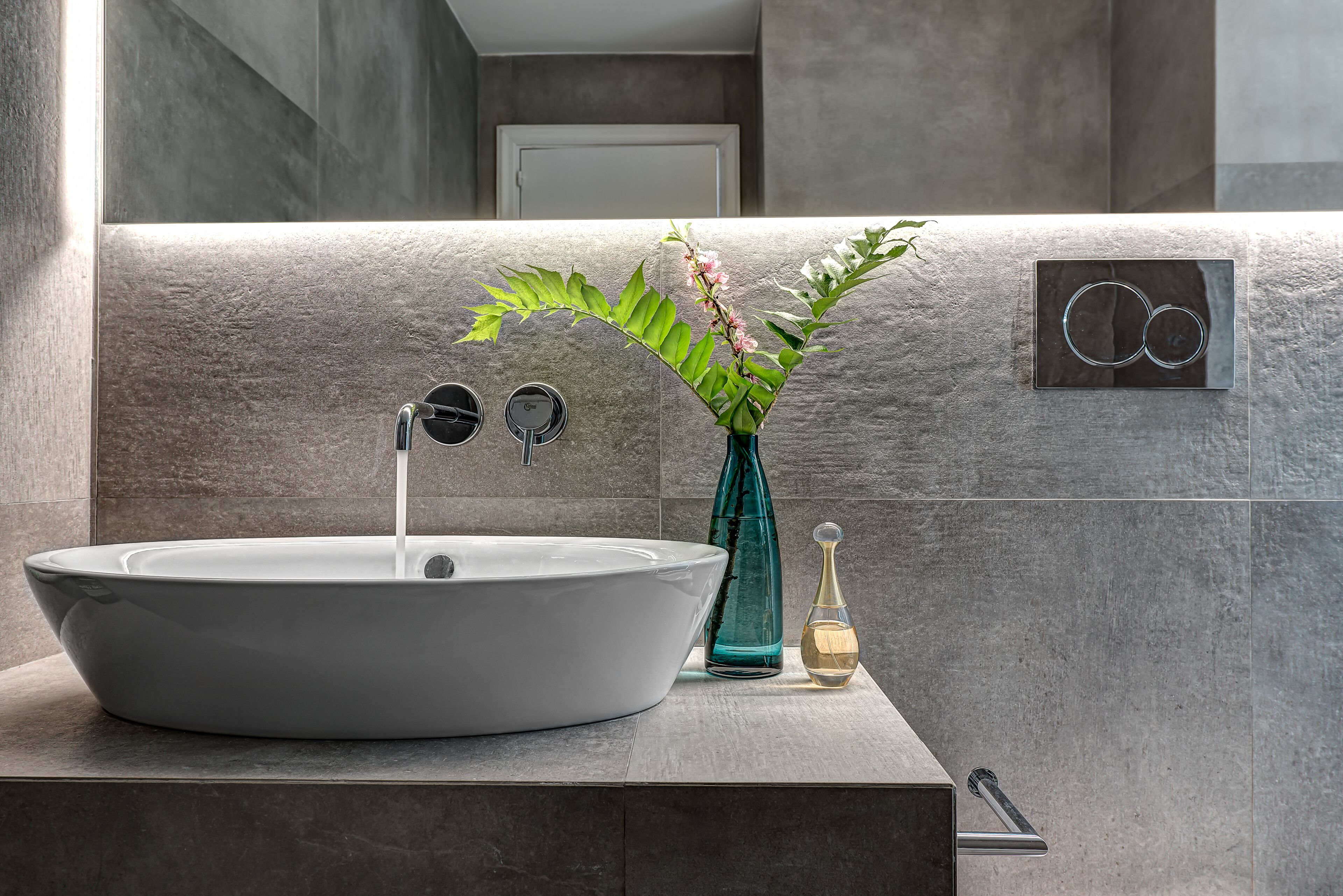 Un baño moderno con un lavamanos grande tipo recipiente, un florero verde con flores y un dispensador de jabón.