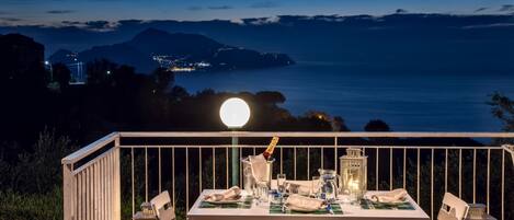 Un área de comedor al aire libre con una mesa dispuesta para dos, con vistas a un paisaje costero nocturno.