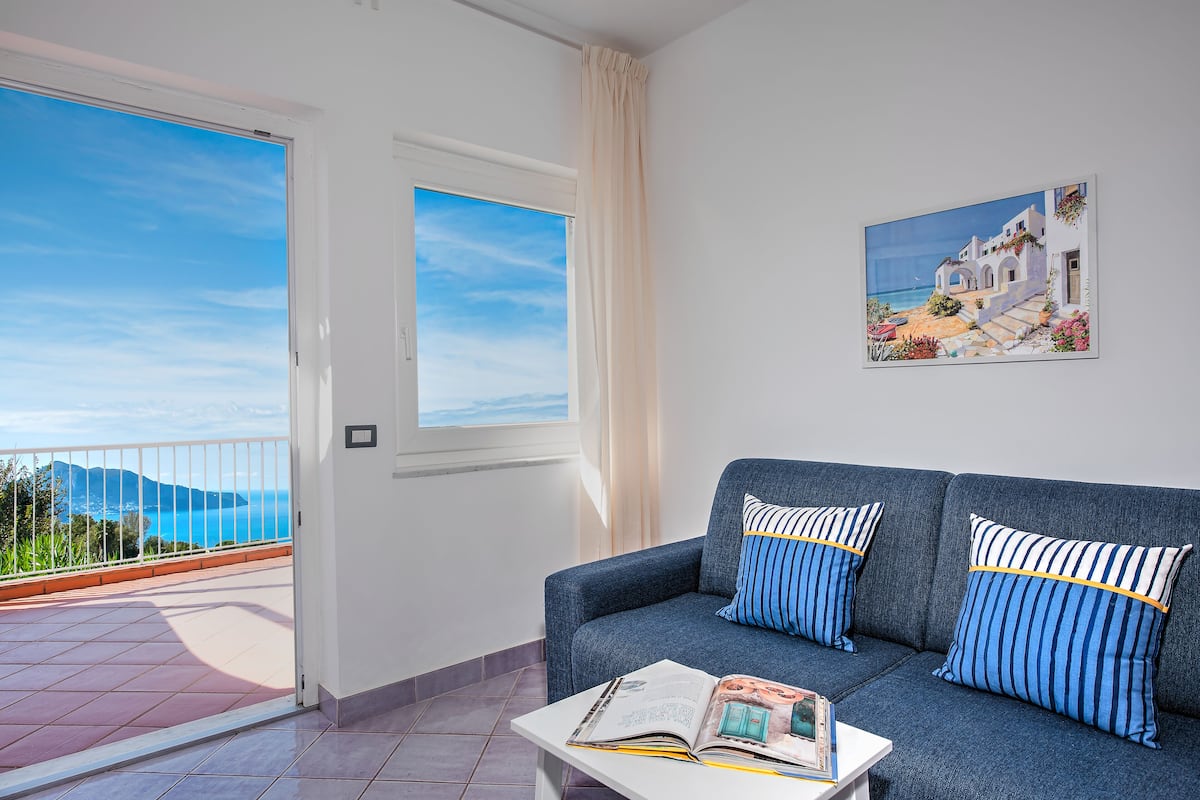Ein Zimmer mit einem blauen Sofa, einem Fenster mit Meerblick und einem Balkon mit Geländer.