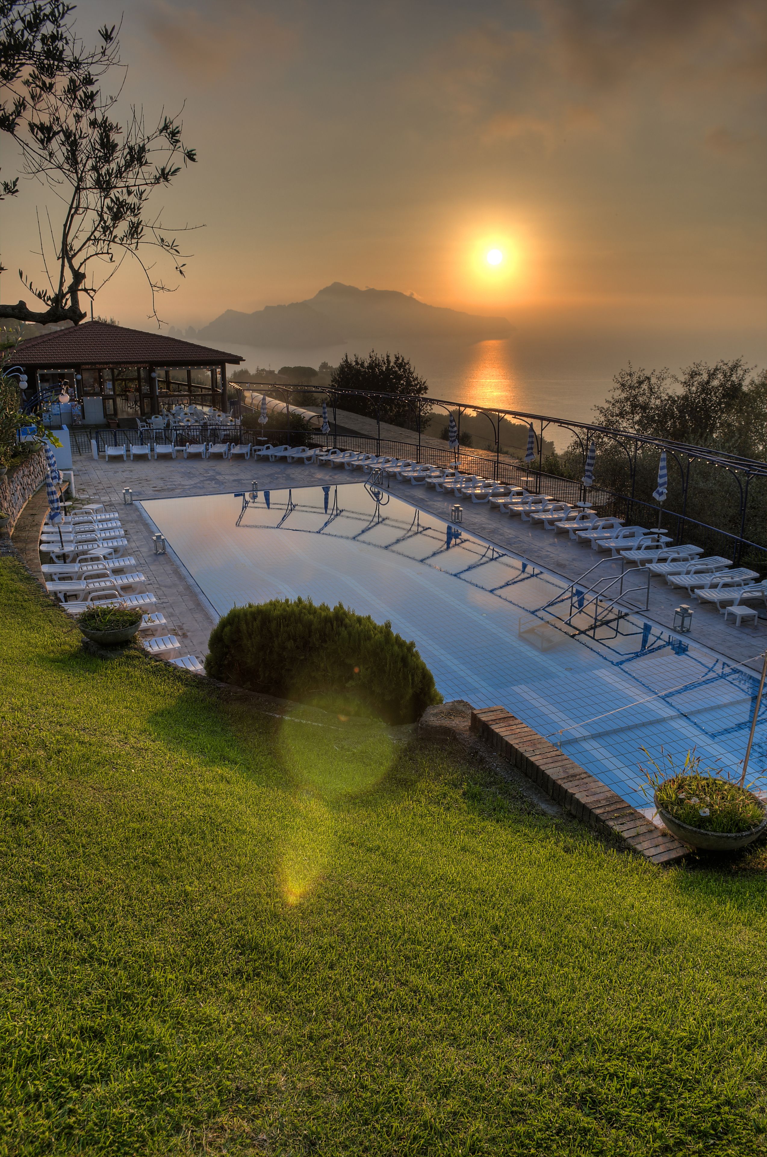 Una piscina con sillones de descanso y un pabellón, rodeada de césped y con vistas a una masa de agua con montañas al fondo.