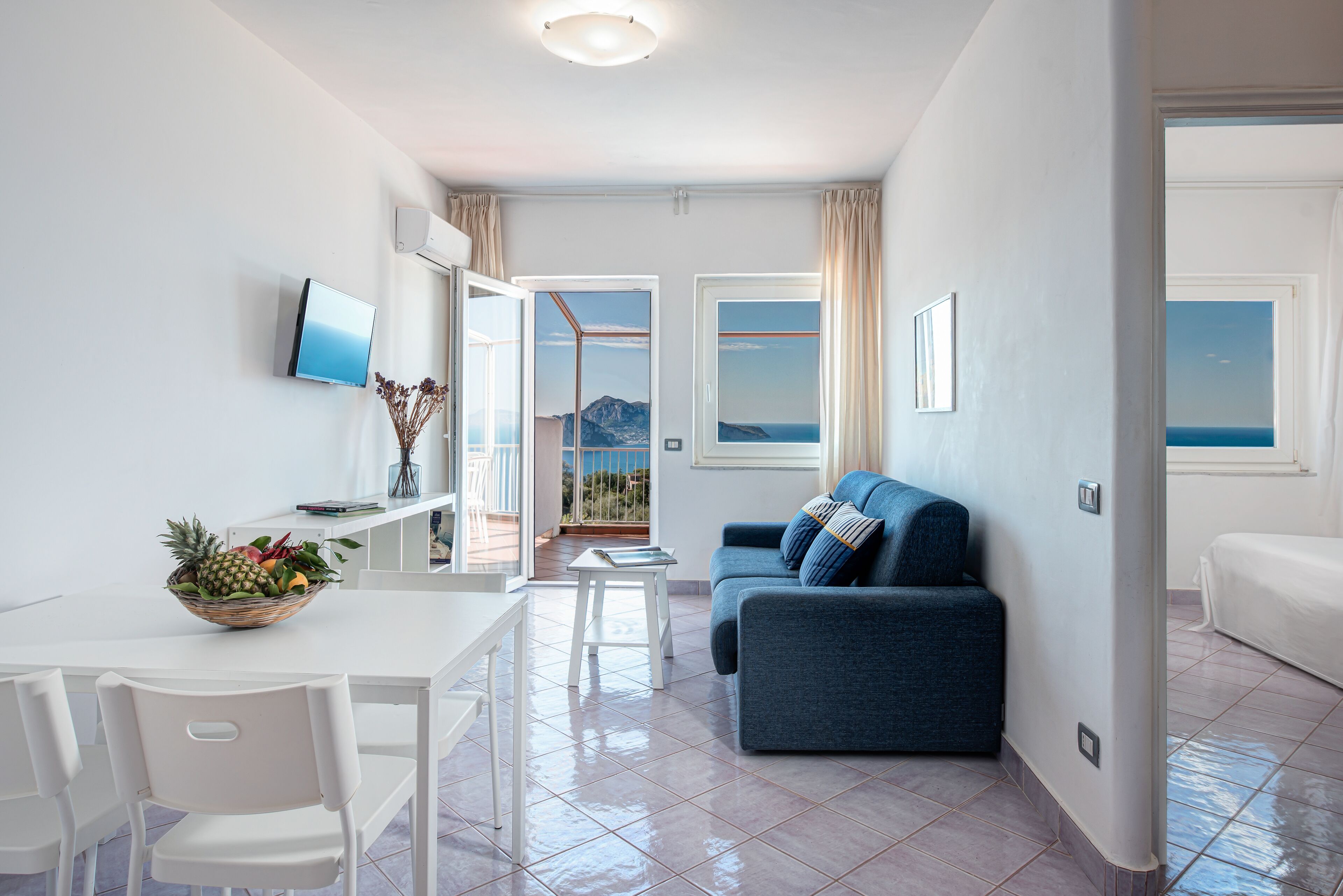 A modern living room with a sofa, a dining table with chairs, a TV mounted on the wall, and a view of the sea through large windows.