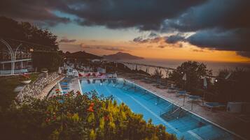 Une piscine entourée de verdure, avec des chaises longues et des parasols, offrant une vue sur la mer au coucher du soleil.