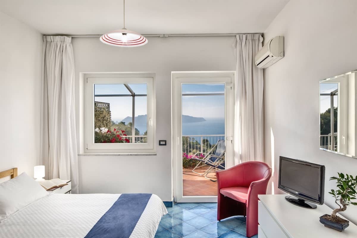 Ein Hotelzimmer mit Bett, Schreibtisch mit Fernseher, einem roten Sessel, einem Balkon mit Aussicht und einer Klimaanlage.