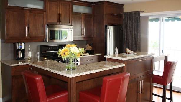 Una cocina con armarios de madera, un refrigerador de acero inoxidable, un microondas y una cafetera. Hay dos taburetes rojos y un florero con flores en la encimera.