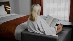 Una persona sentada en un sofá gris en una habitación de hotel, leyendo un libro.