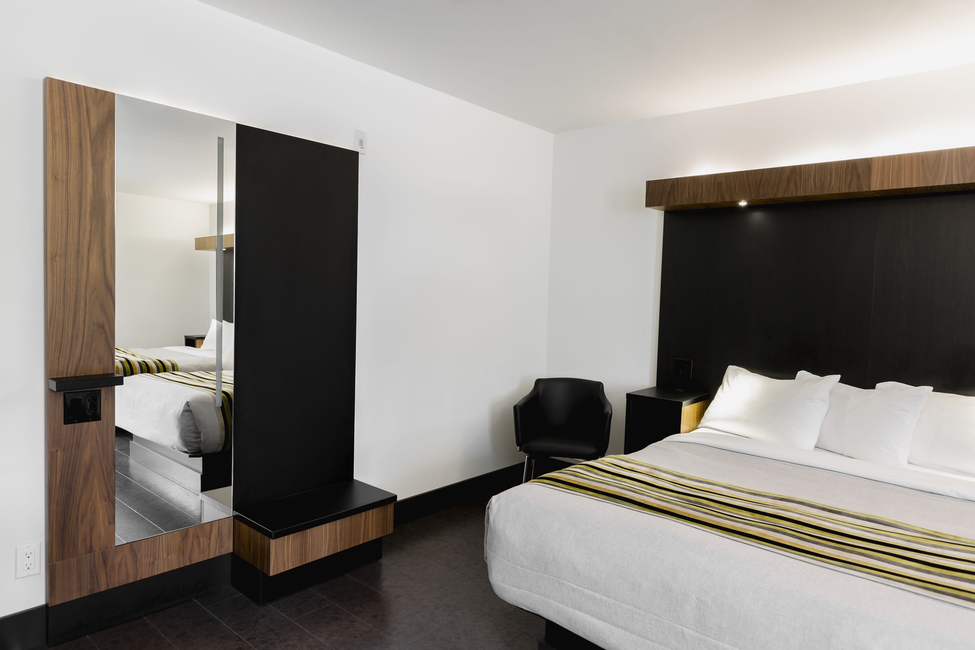 A modern hotel room with a large bed, a black chair, a nightstand, and a mirror.