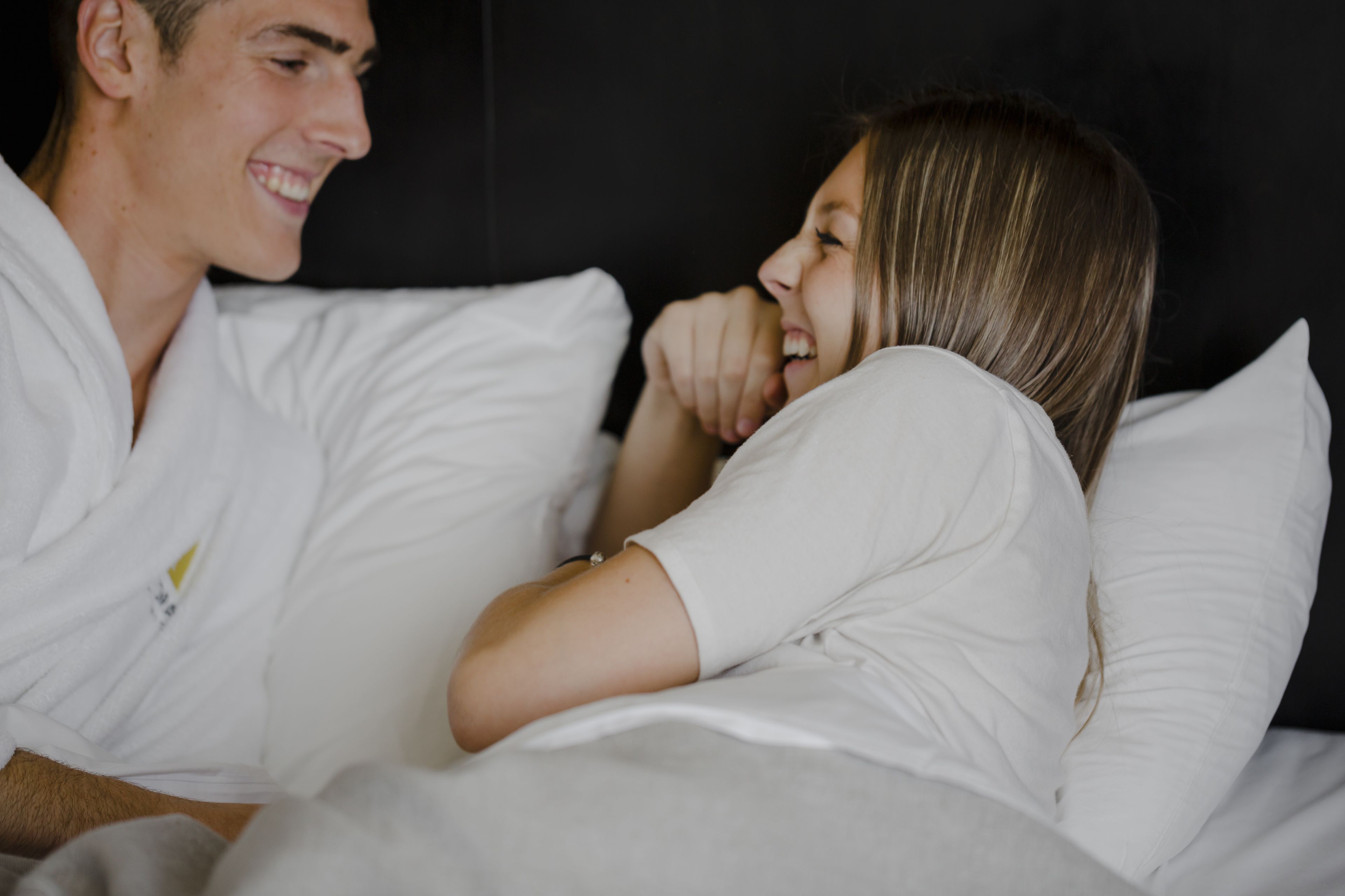 Um casal na cama com roupa de cama e almofadas brancas, num quarto com parede escura.