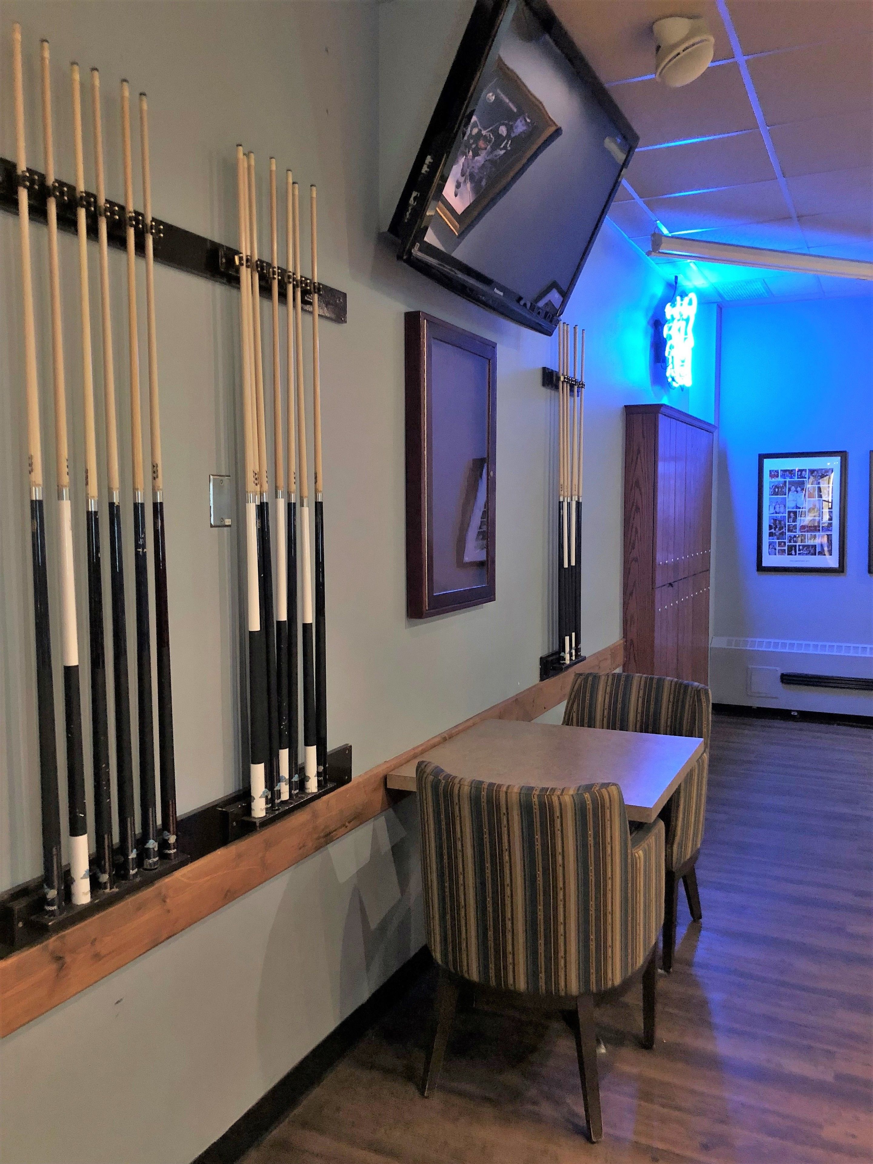 A billiards room with a long table and striped chairs, a wall-mounted pool table, and a neon sign.