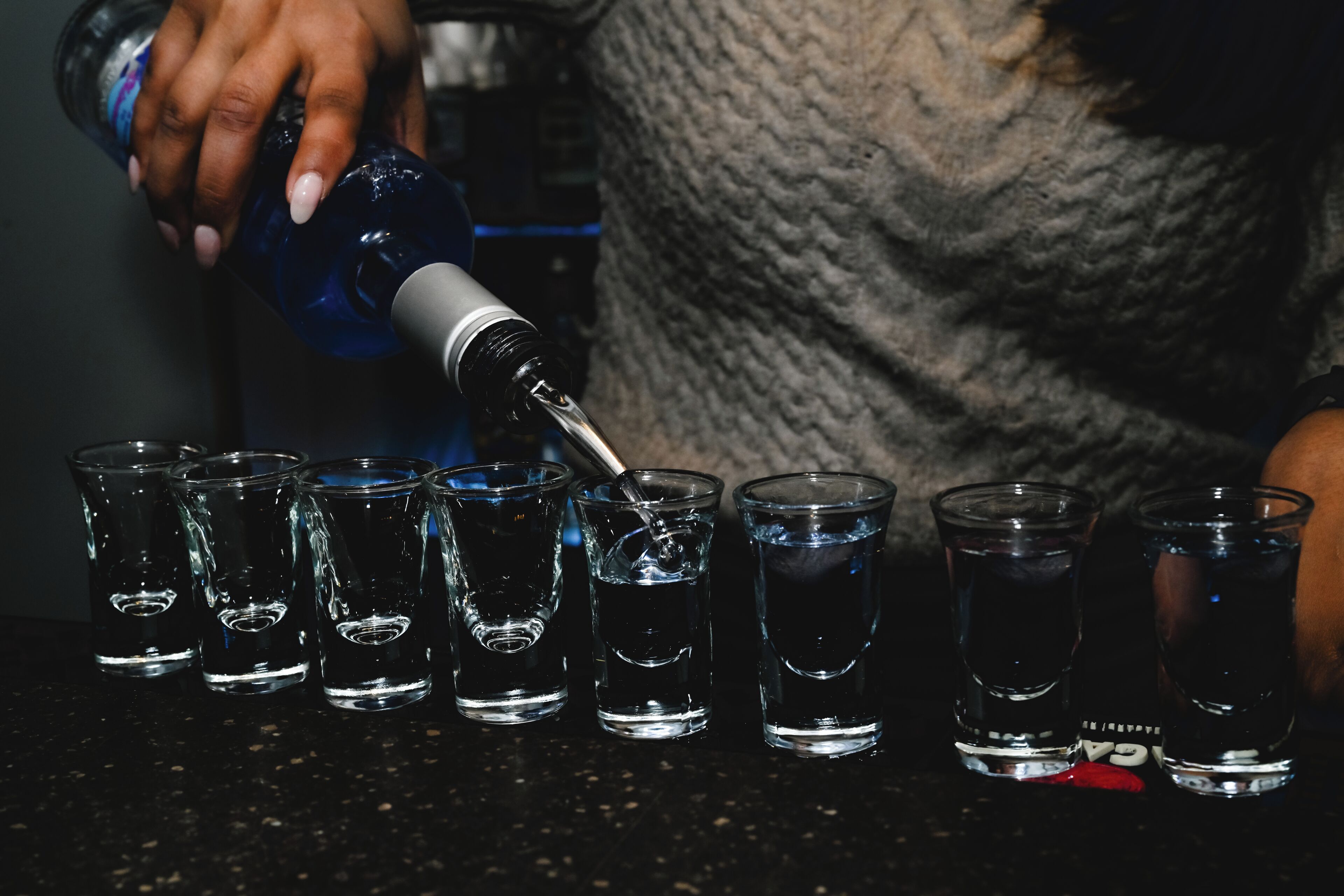 A person pouring a dark liquid into shot glasses on a bar counter.
