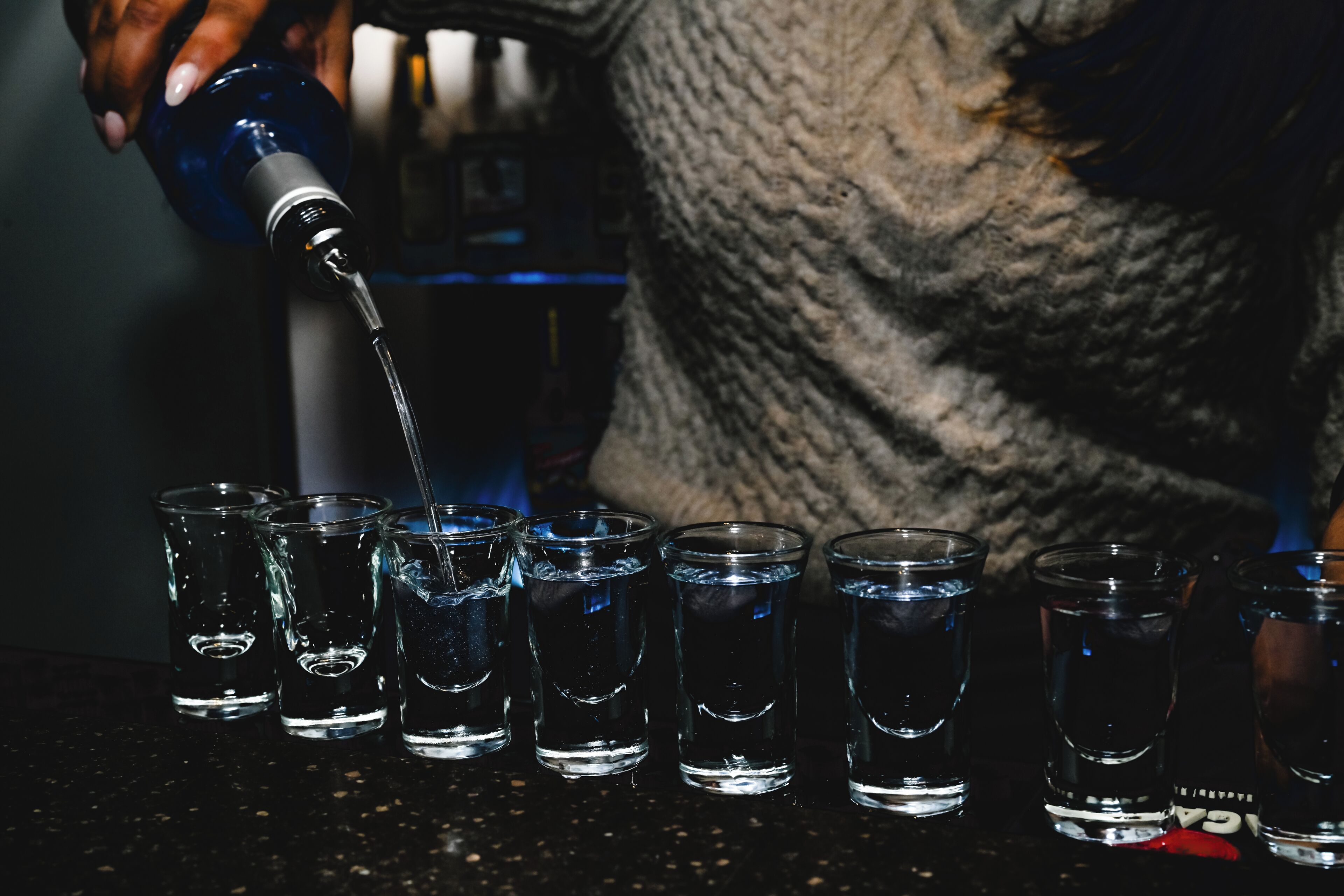 Une personne verse un liquide transparent dans des petits verres à shooter sur le comptoir d’un bar.