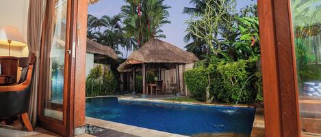 A poolside view with a glass door leading to a room, a thatched-roof structure, and lush greenery.