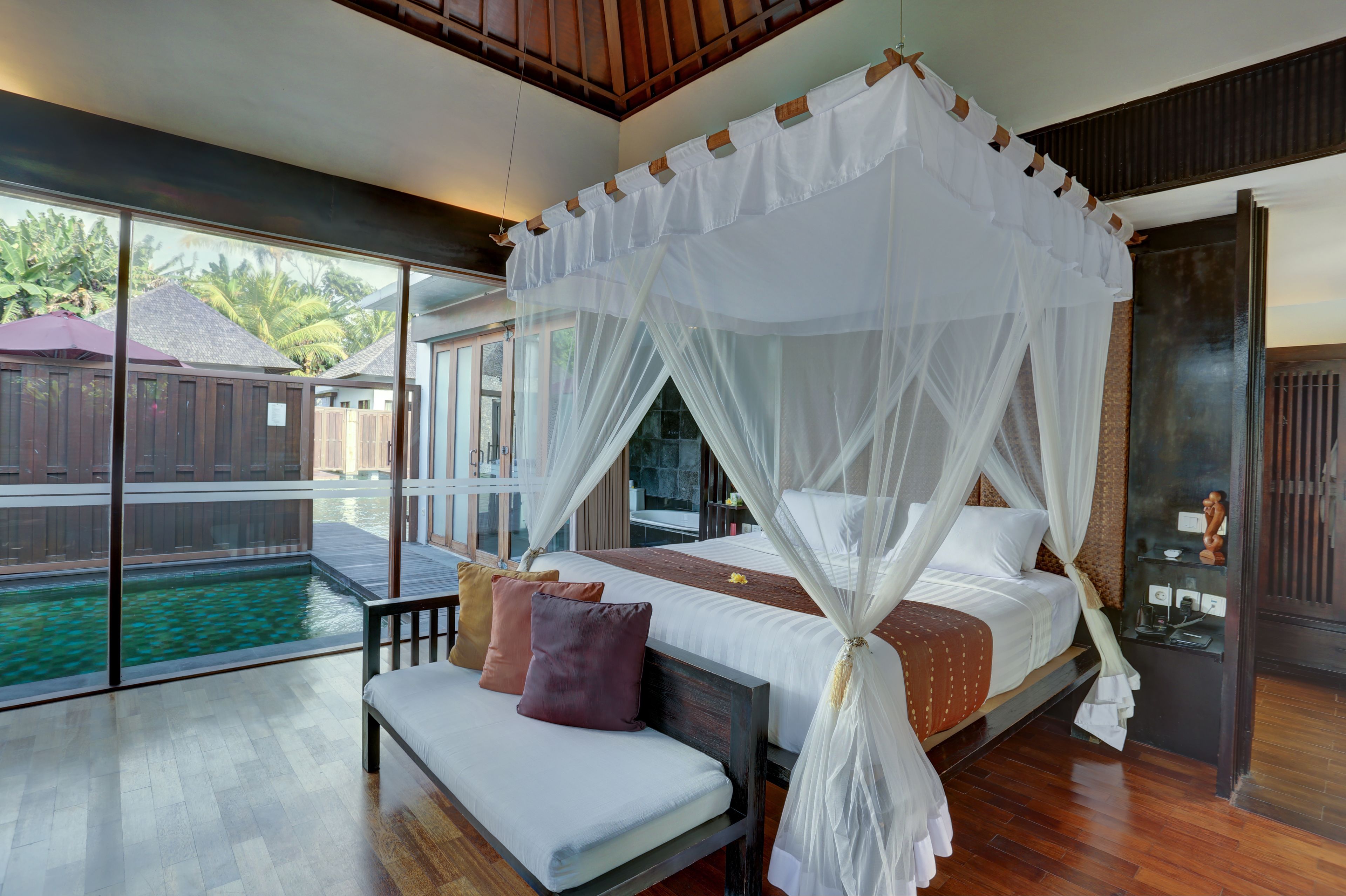 A bedroom with a four-poster bed, a bench, and a view of a pool.