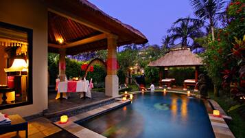 A poolside area with a covered dining area, a thatched-roof structure, and a well-lit evening setting.