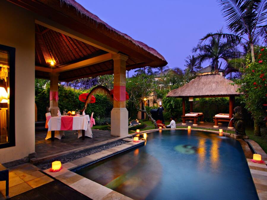 A poolside area with a covered dining area, a thatched-roof structure, and a well-lit evening setting.