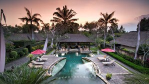 A resort pool area with lounge chairs and umbrellas, surrounded by palm trees and thatched-roof huts. - FuramaXclusive Resort & Villas, Ubud (Mas)
