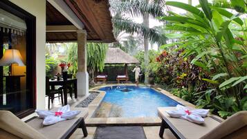 A pool area with lounge chairs and a table, surrounded by tropical plants and a thatched-roof structure.