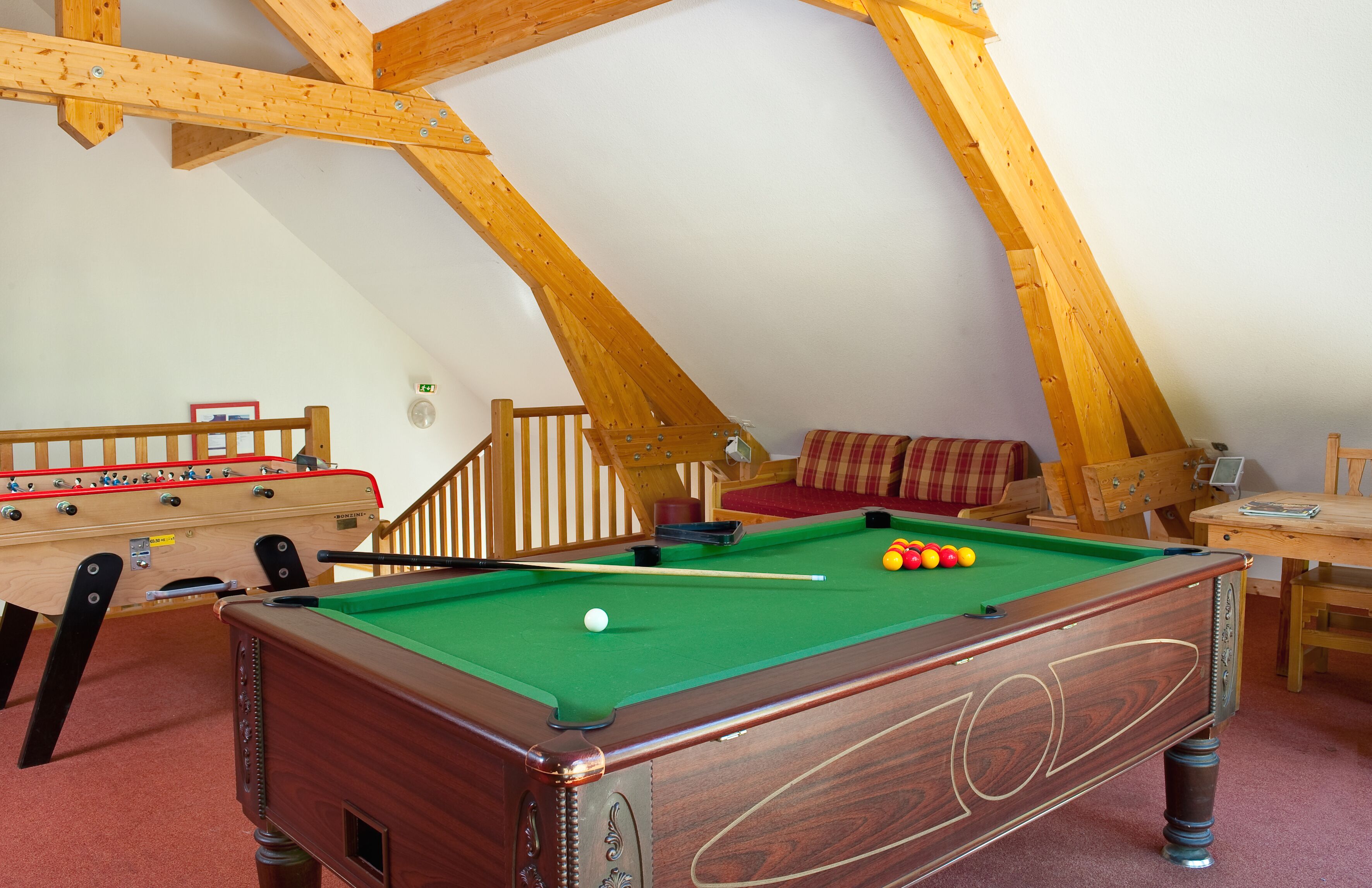 A pool table with balls, a foosball table, and a sofa in a room with wooden beams.
