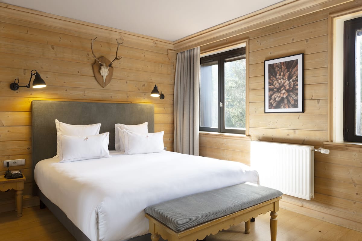 A wooden-paneled room with a large bed, two bedside lamps, a mounted deer skull, a framed picture, and a window with curtains.