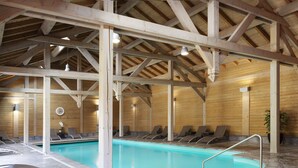 An indoor swimming pool with wooden beams, lounge chairs, and a tiled floor.