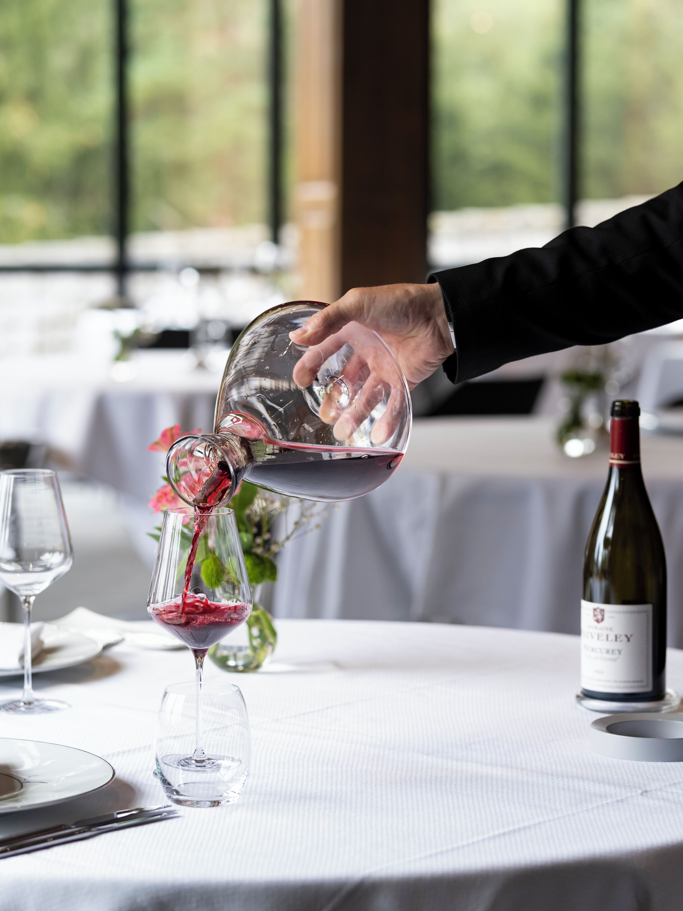 Una mano sirviendo vino tinto en una copa sobre una mesa, con una botella de vino y un jarrón de flores al fondo.