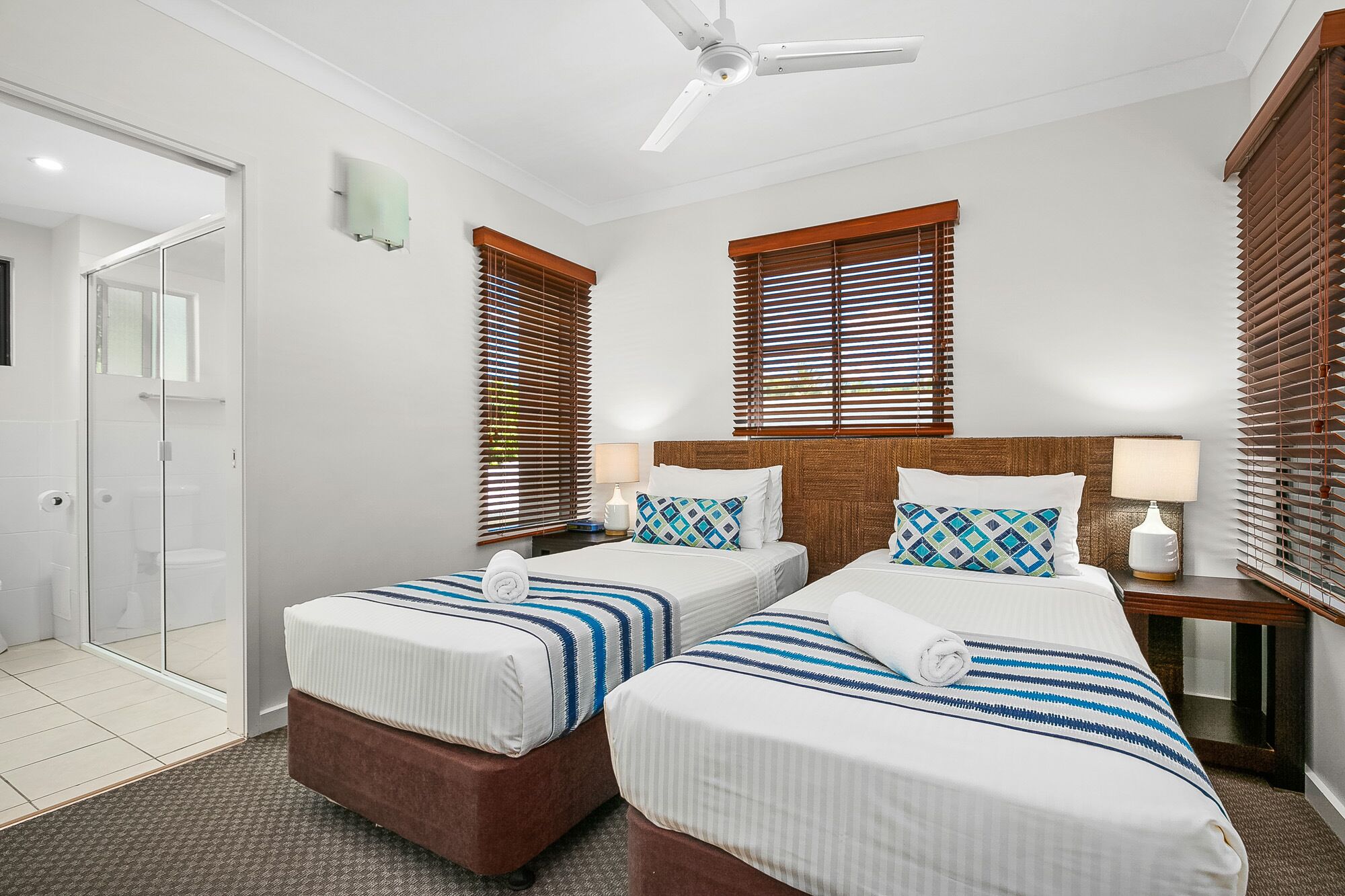 Habitación de hotel con dos camas, ventilador de techo y persianas de madera en las ventanas.