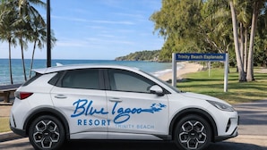 A white SUV with "Blue Lagoon Resort Trinity Beach" branding parked on a road with a beach and palm trees in the background. - Blue Lagoon Resort (Trinity Beach)