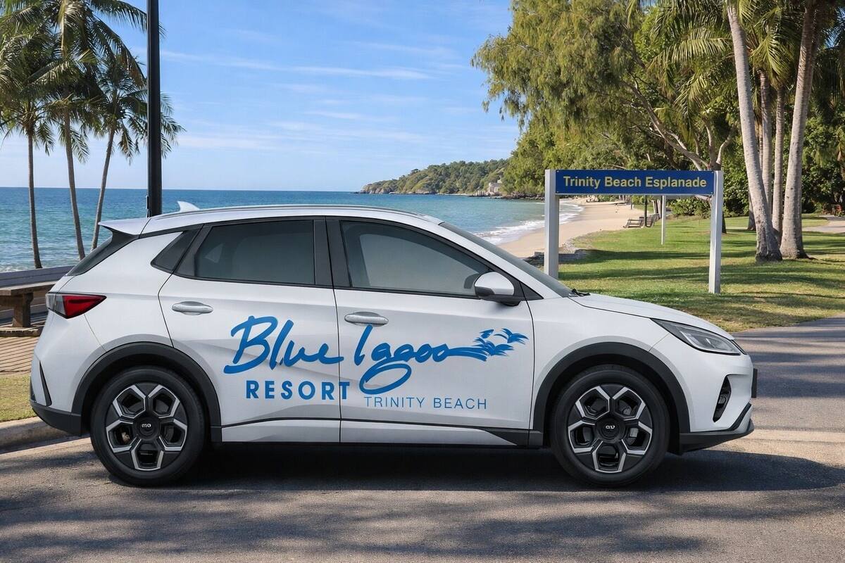 Ein weißer SUV mit der Aufschrift "Blue Lagoon Resort Trinity Beach" auf einer Straße, im Hintergrund ein Strand und Palmen.