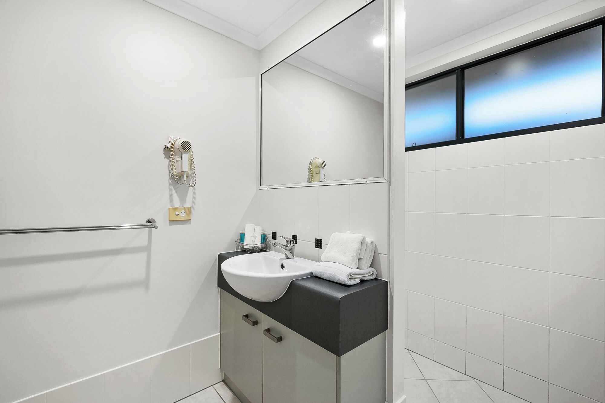 Un baño moderno con un lavabo blanco, encimera negra y un espejo encima. Hay un perchero y un secador de pelo en la pared.
