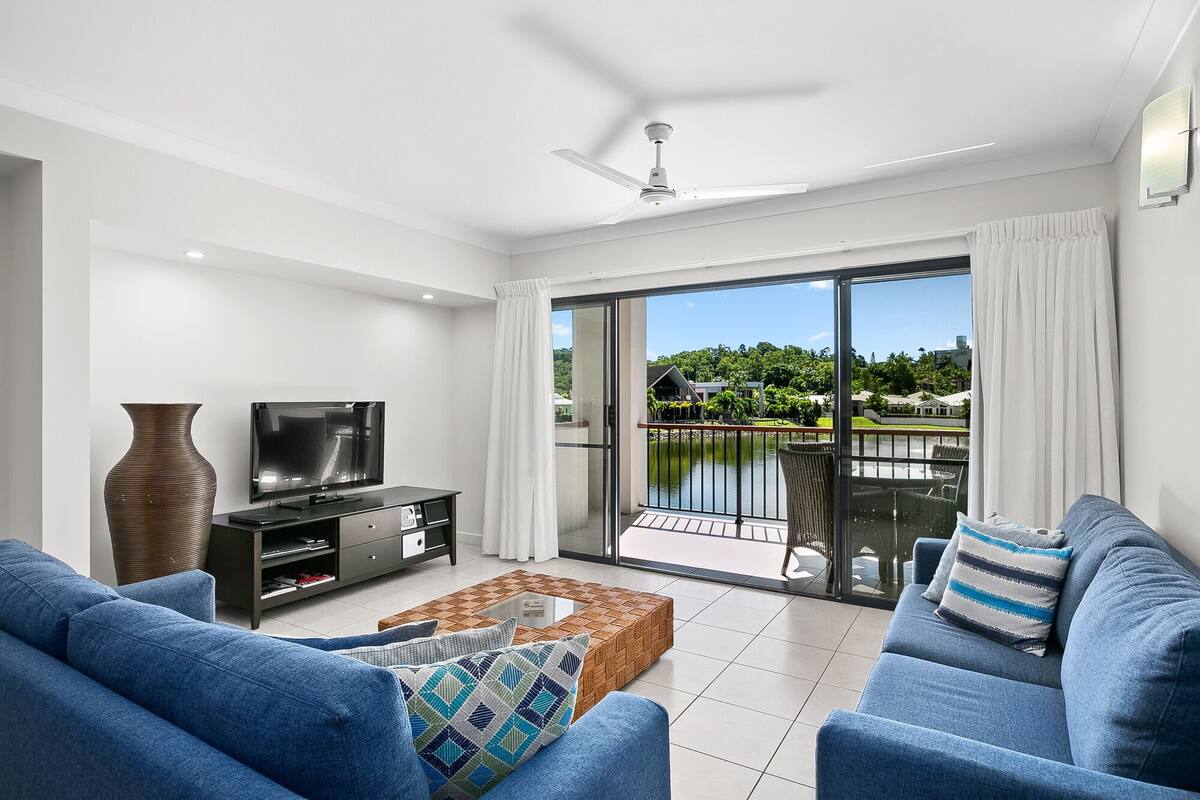 Ein modernes Wohnzimmer mit Fernseher, Sofa und Balkon mit Blick auf einen See und viel Grün.