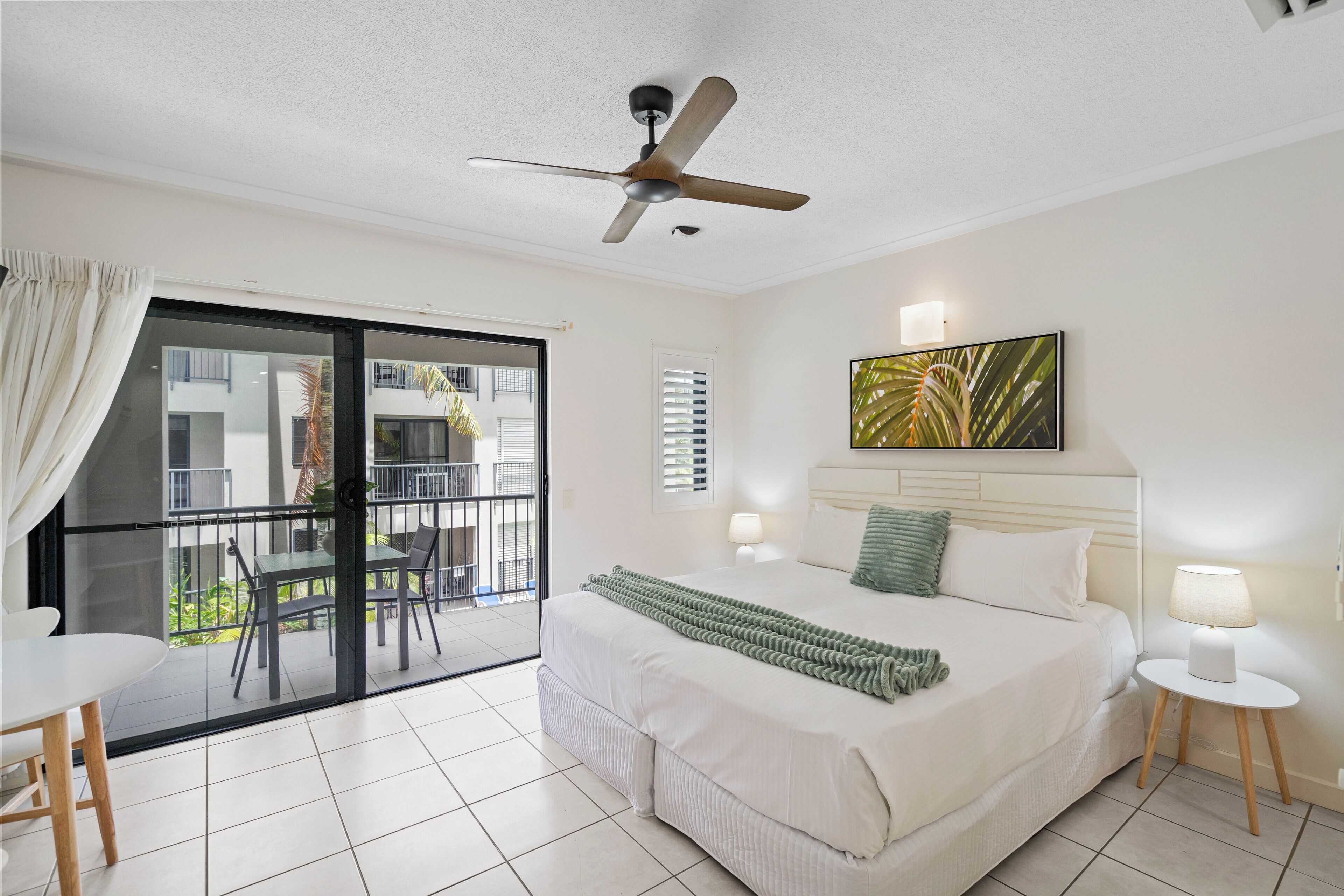A hotel room with a bed, a ceiling fan, a balcony with a table and chairs, and a view of buildings outside.