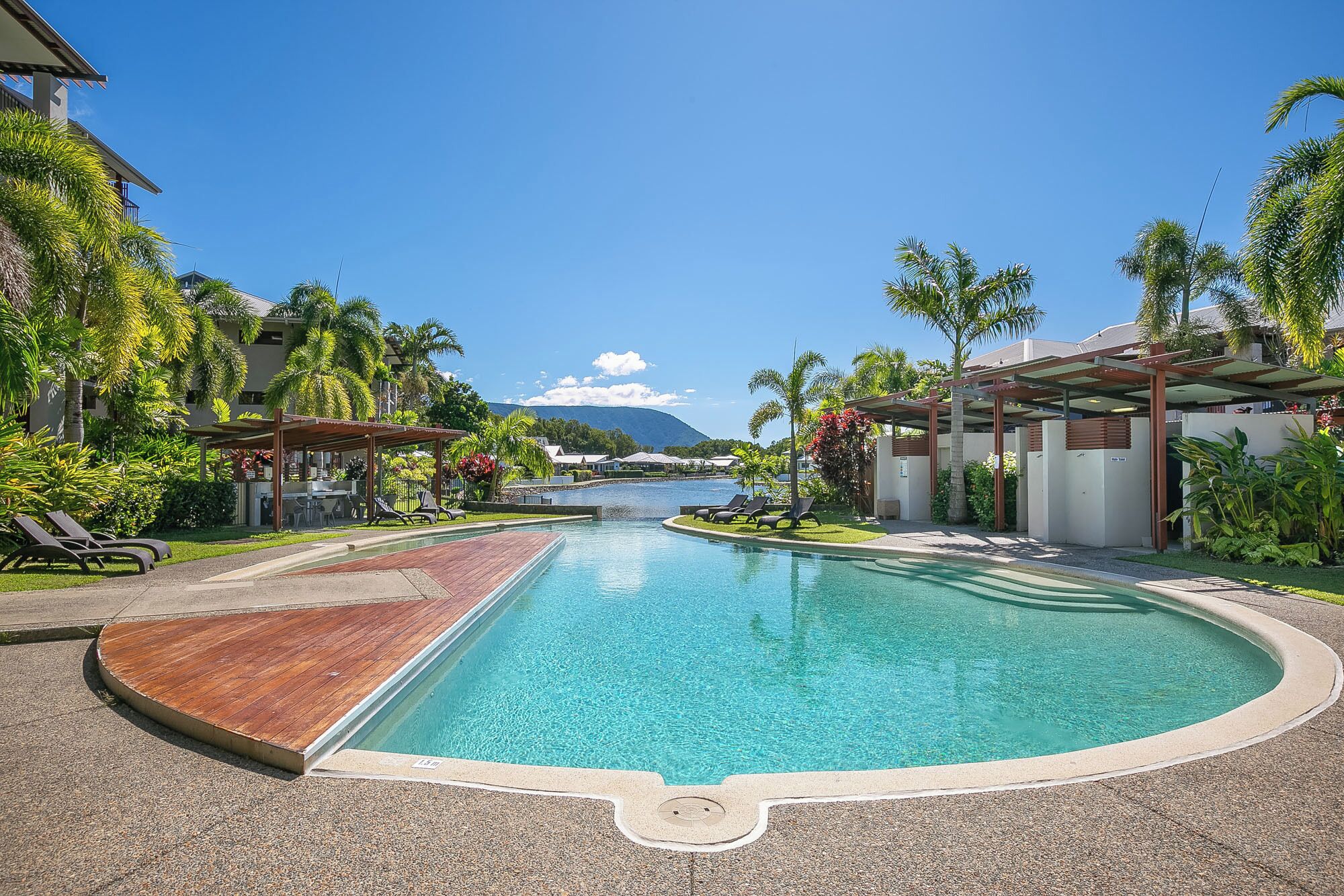 A resort pool with a wooden deck, lounge chairs, and palm trees.