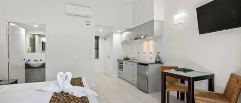 Una habitación de hotel con cama, una mesa de comedor con sillas, una zona de cocina y un televisor montado en la pared.