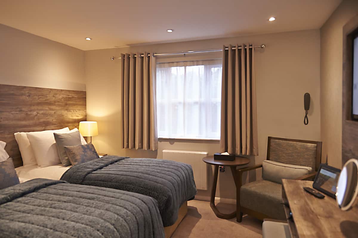 Une chambre d’hôtel avec deux lits, un bureau, une chaise, une lampe et une fenêtre avec des rideaux.