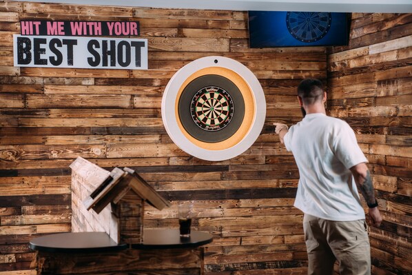 A dartboard mounted on a wooden wall with a sign above it that reads "HIT ME WITH YOUR BEST SHOT." - Drift Inn at Wyboston Lakes (Bedford)