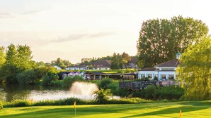 A golf course with a pond, a wooden bridge, and houses in the background. - Drift Inn at Wyboston Lakes (Bedford)