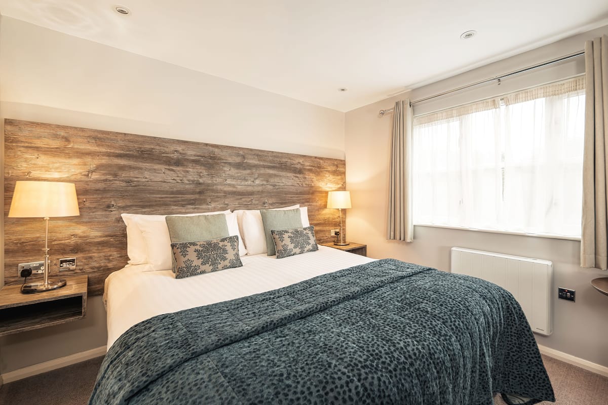 Une chambre d’hôtel avec une tête de lit en bois, un grand lit recouvert d’une couverture bleue, deux lampes de chevet et une fenêtre avec des rideaux.