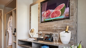 A hotel room with a TV displaying a watermelon ad, a minibar, a mirror, a bedside table with a bottle of champagne, a robe, and a flower arrangement. - Drift Inn at Wyboston Lakes (Bedford)