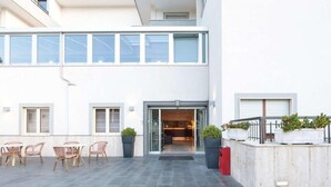 Un edificio moderno con fachada plana, amplios ventanales y una zona de estar al aire libre cubierta con sillas y plantas en macetas.