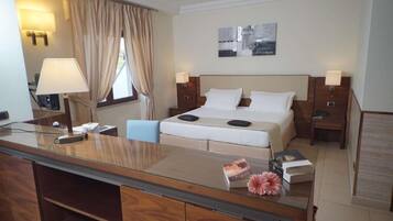 A hotel room with a bed, bedside tables, a desk with a lamp, a chair, and a window with curtains.