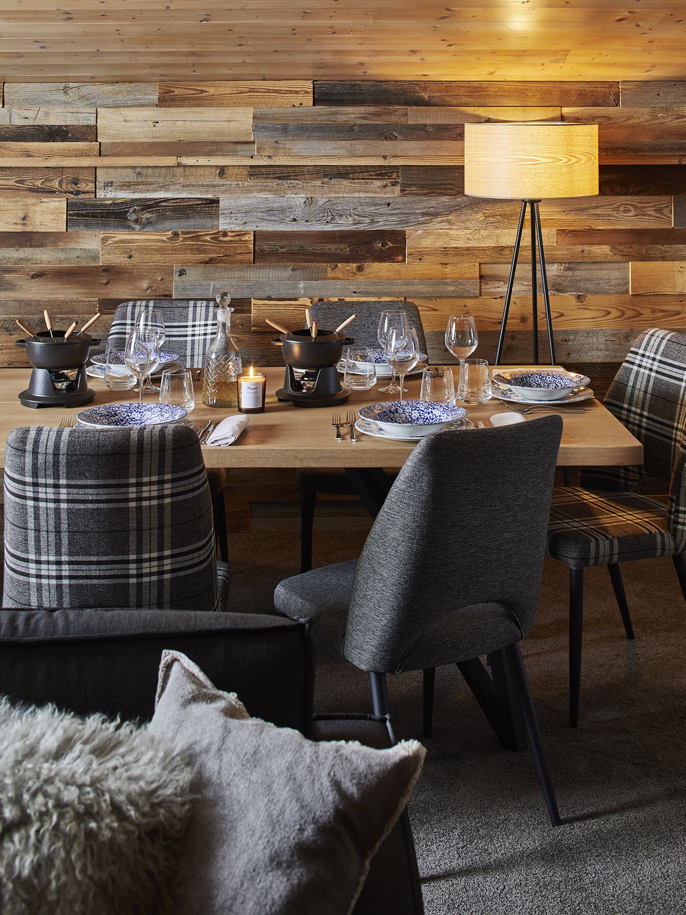 A dining area with a wooden wall backdrop, a table set for four with chairs, a floor lamp, and a candle.