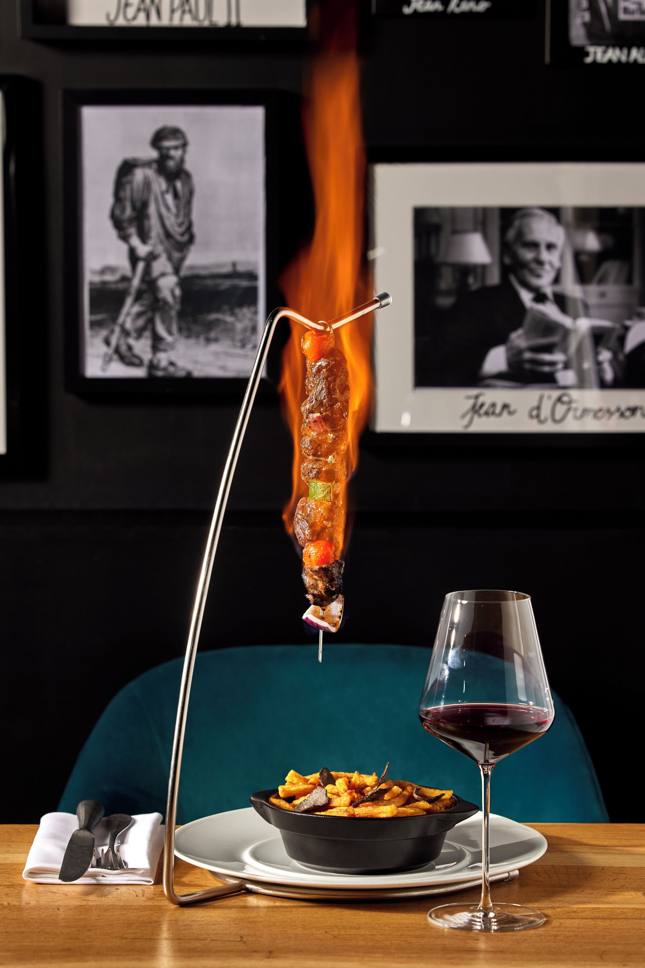 A dining table with a plate of food, a glass of red wine, and a skewer of meat over a flame.